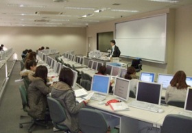 先生がホワイトボードの前に立ち、パソコンの前に座る生徒たちに授業をしている様子
